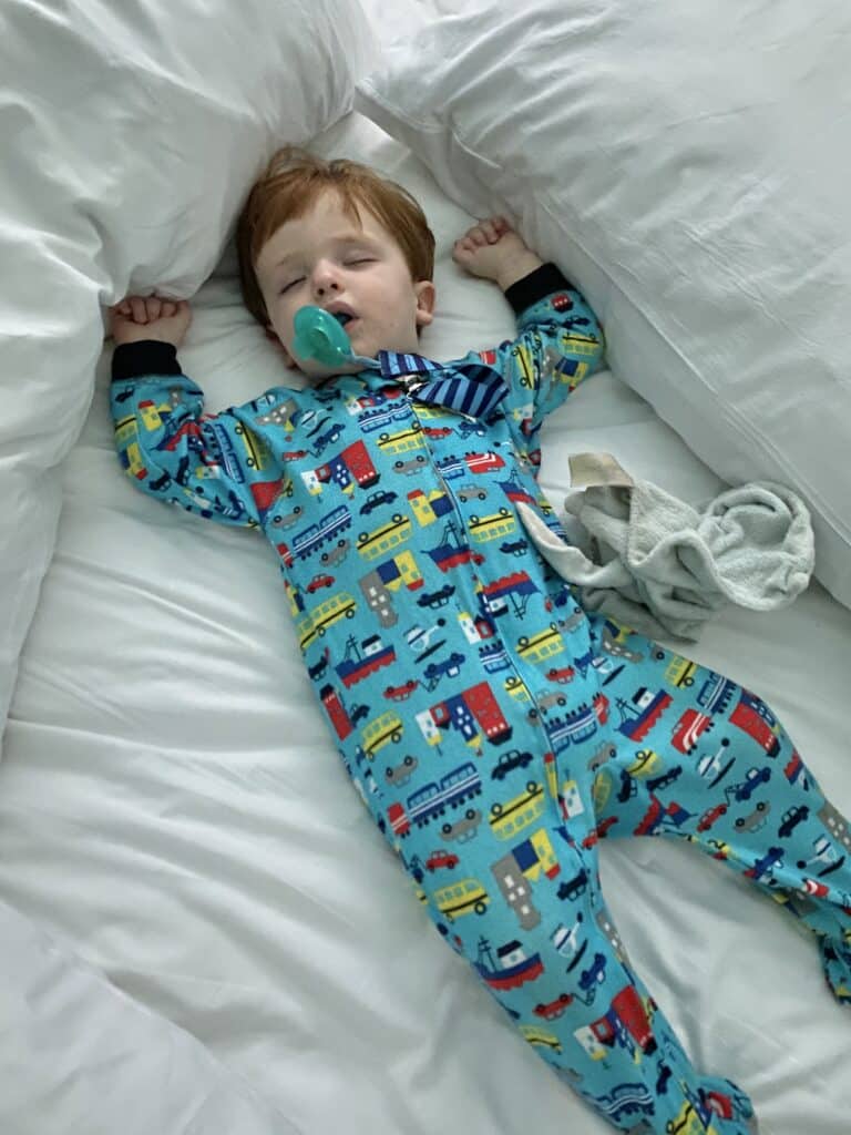 Baby in blue vehicle pajamas sleeping in a bed with a pacifier in his mouth